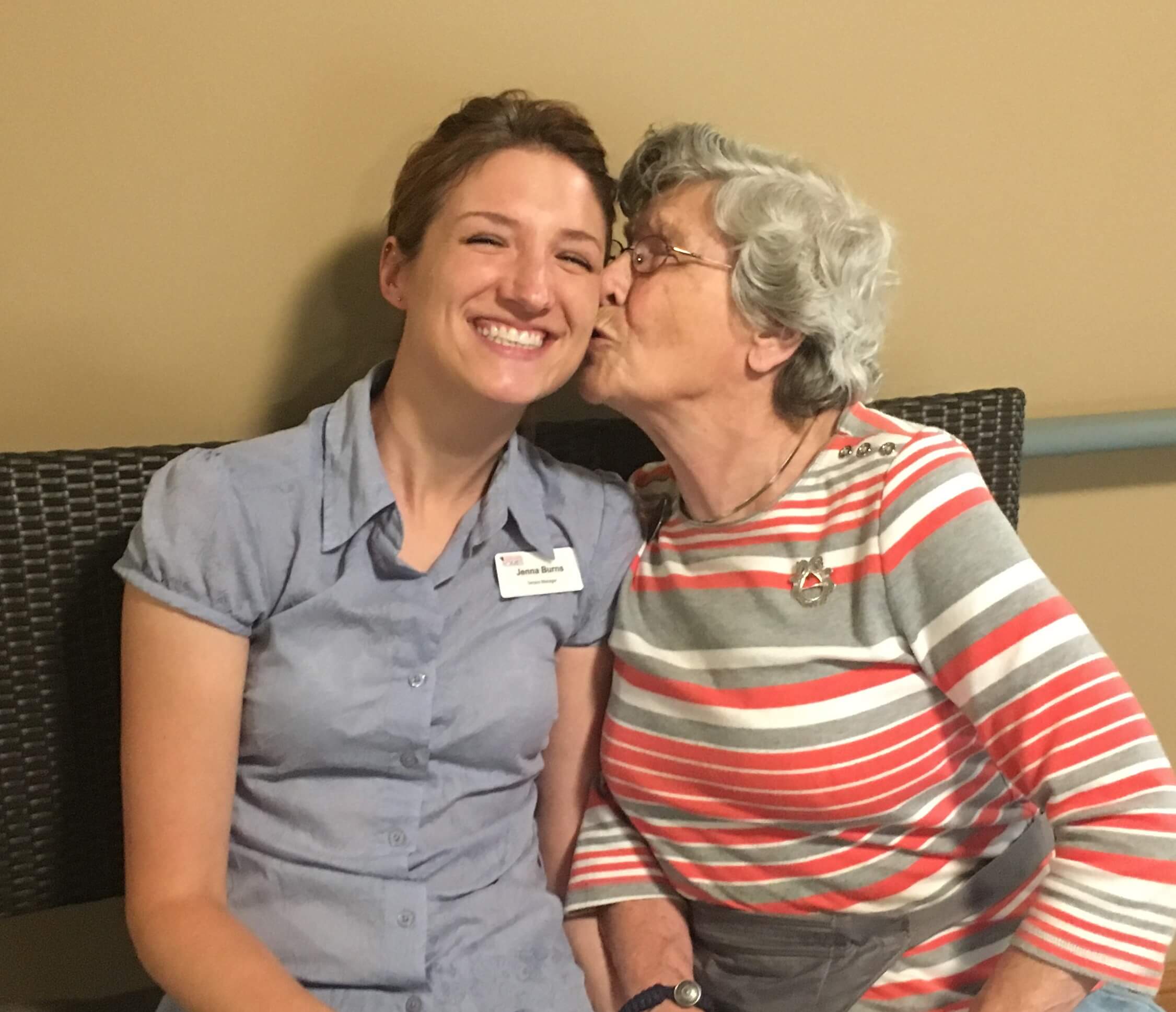 KSU alumna Jenna Kilawee smiles great big while holding hands with an older woman who's kissing her on the cheek.  They are seated on a bench in the facility where the young woman works and the older woman lives.